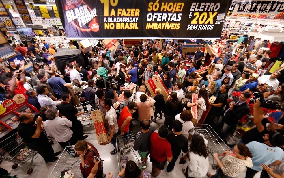 Multidão de consumidores em uma loja lotada, com carrinhos e caixas de produtos, sob um letreiro de Black Friday