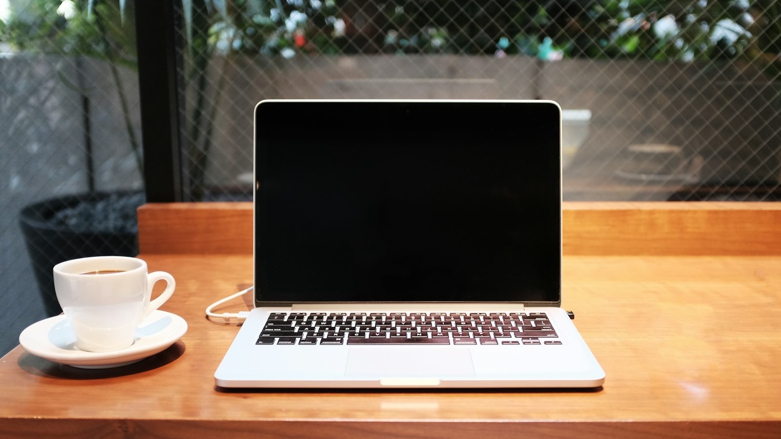 Notebook aberto com a tela apagada sobre uma mesa de madeira, ao lado de uma xícara de café
