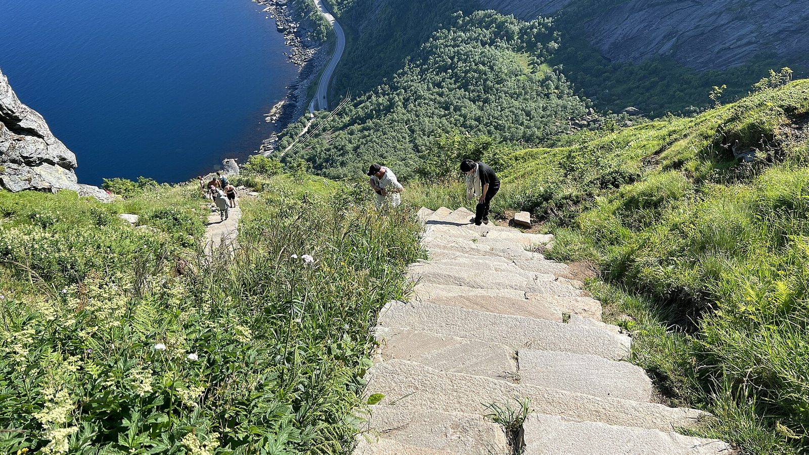 Longa escadaria de pedra subindo uma encosta verde com o mar e uma estrada ao fundo, com algumas pessoas distantes na subida