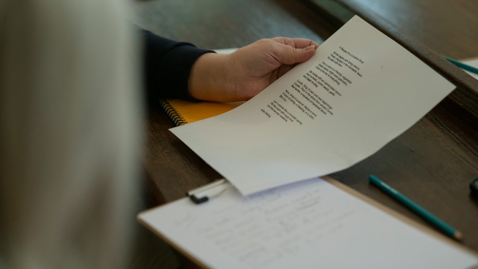 Pessoa segurando uma folha de papel impressa sobre uma mesa de madeira, ao lado de uma prancheta