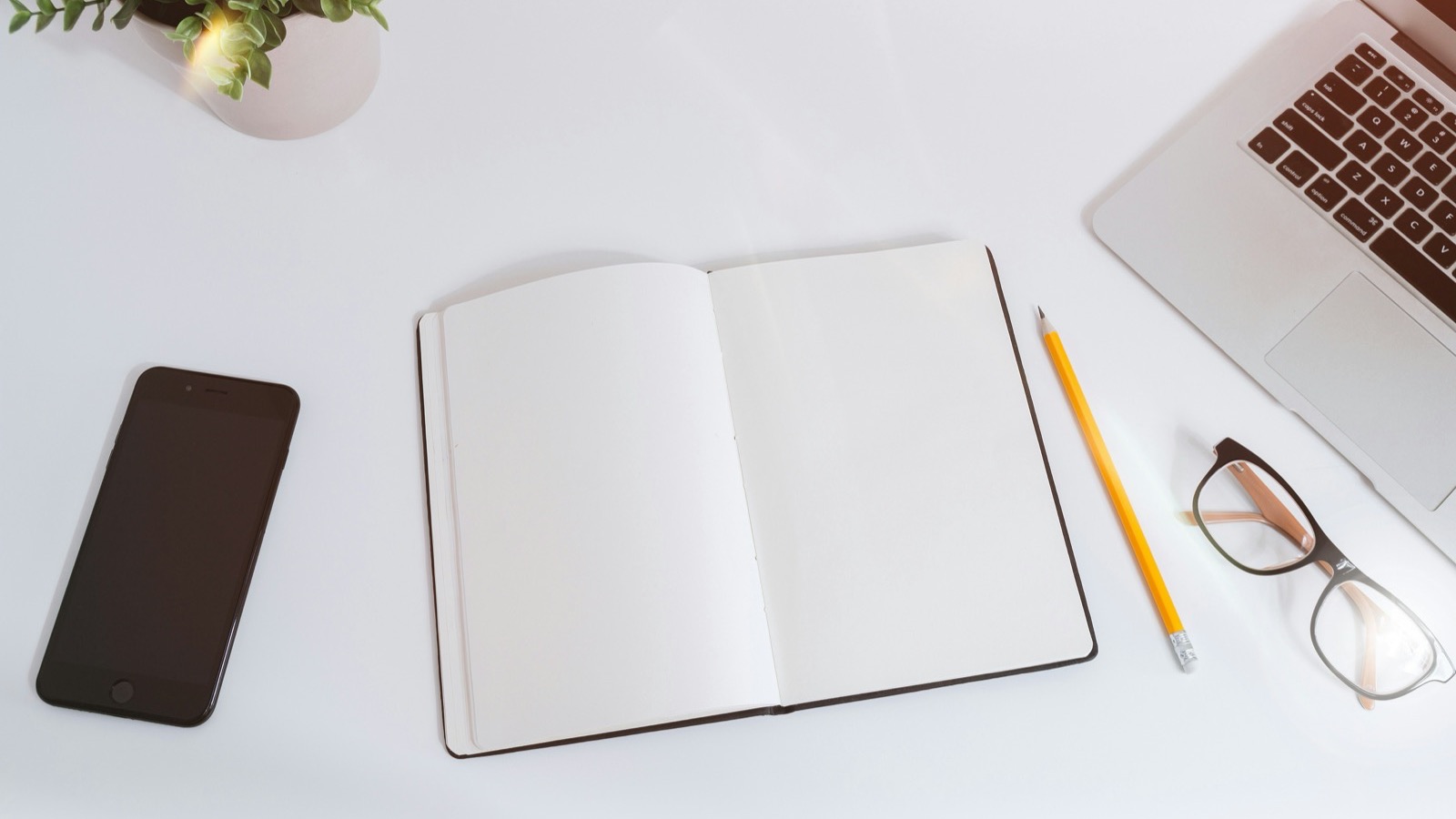Mesa branca com caderno aberto em branco, lápis, óculos, celular e parte de um notebook