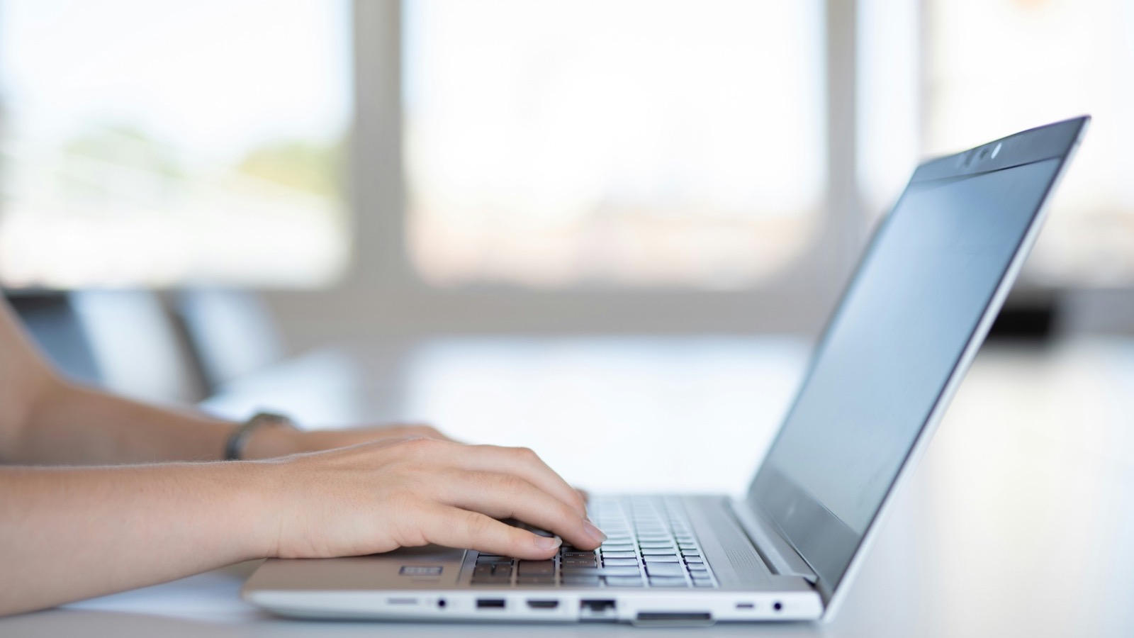 Mãos digitando em um notebook sobre uma mesa clara, em um ambiente com janelas ao fundo