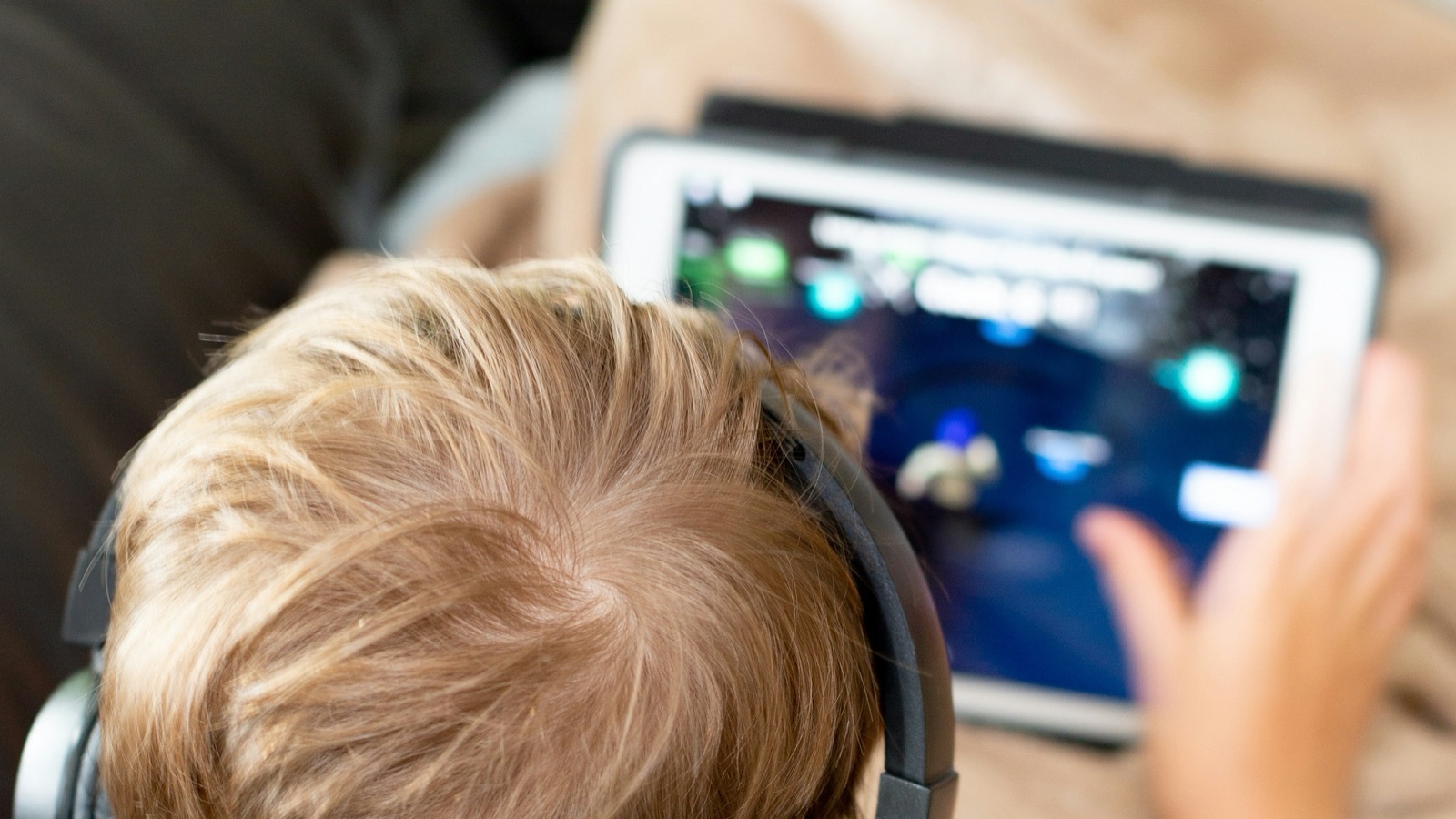 Criança de costas, com fones de ouvido, usando um tablet com um jogo na tela