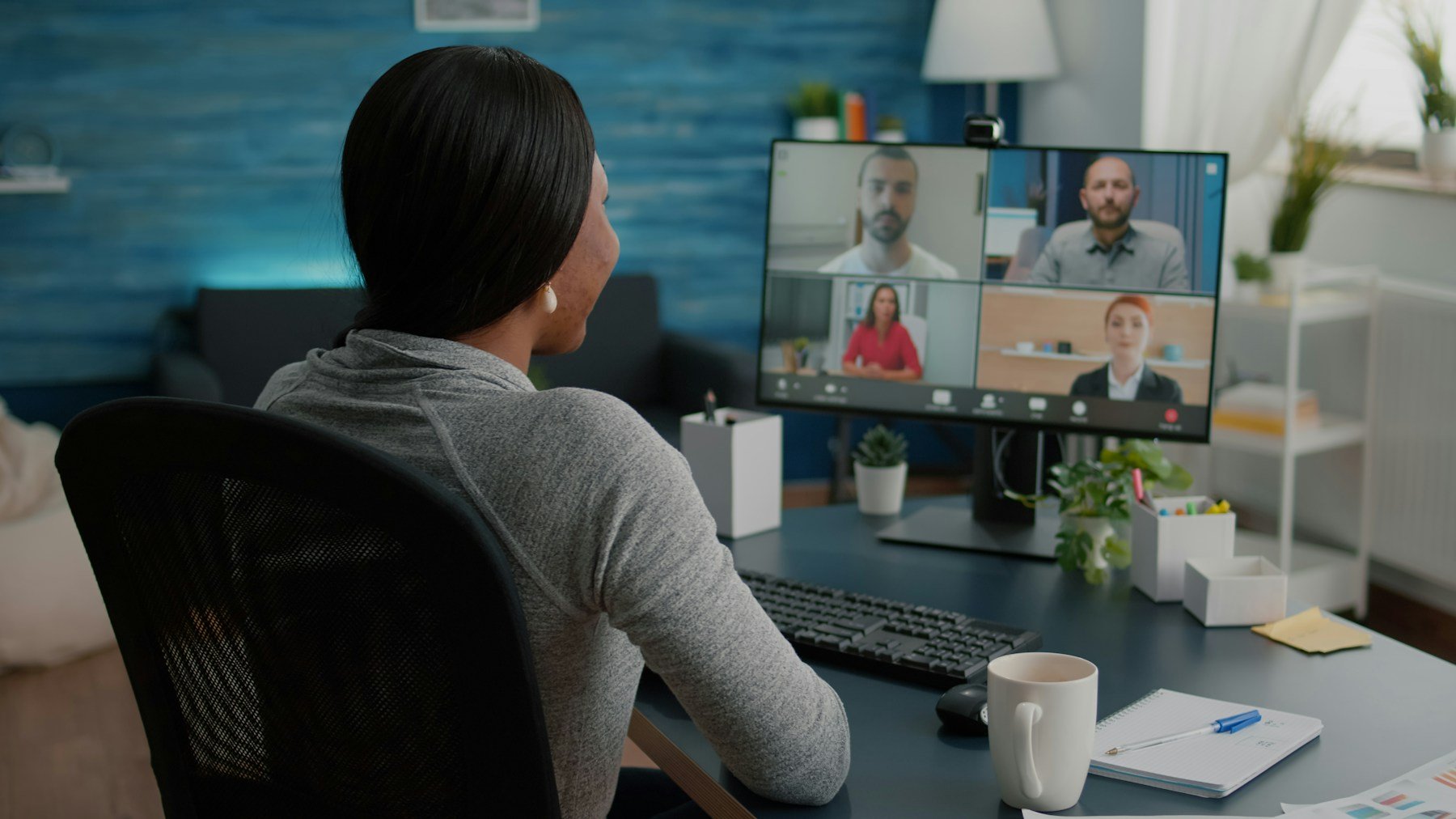 Mulher de costas diante de um monitor com uma chamada de vídeo em quatro quadros, em uma mesa com teclado e caneca
