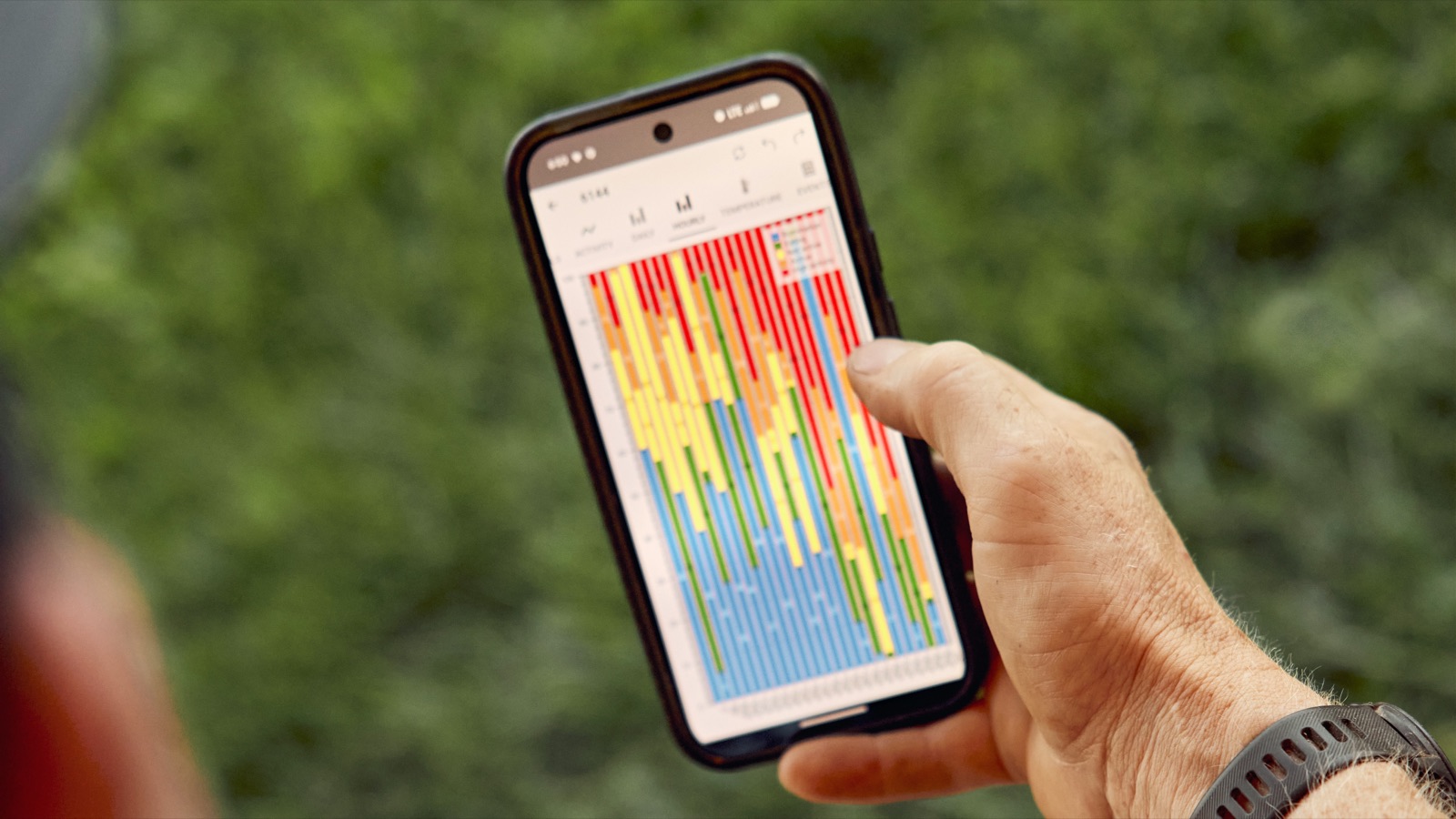 Mão segurando um smartphone com um gráfico de barras coloridas, sobre fundo de vegetação desfocada