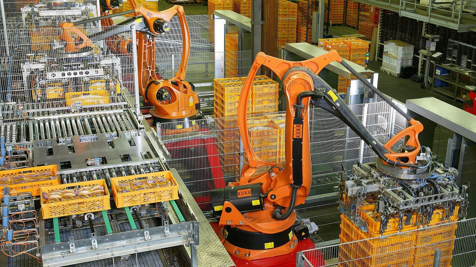 Braços robóticos laranja empilhando caixas de pão amarelas em uma fábrica, dentro de células cercadas por grades